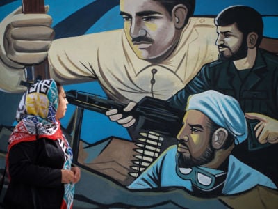 A woman in a hijab looks at a mural depicting several soldiers fighting