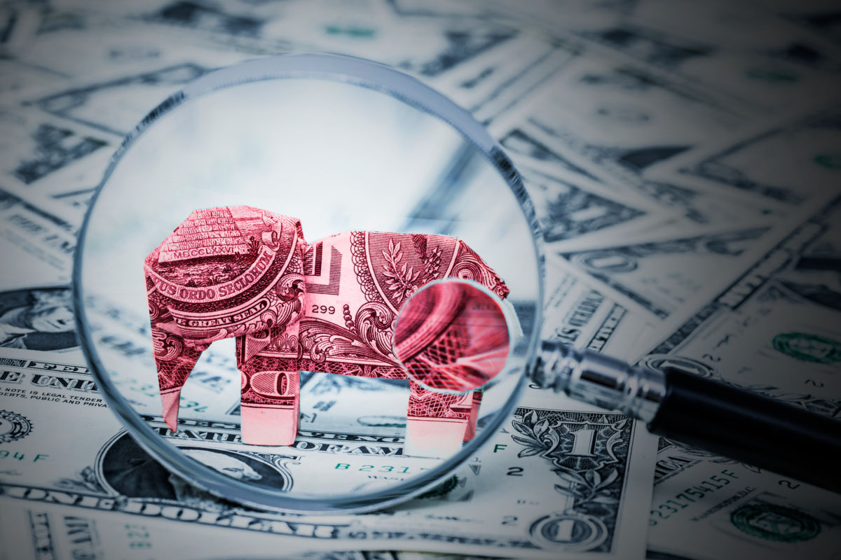 An oragami elephant made of money stands among other us currency while being inspected by a microscope