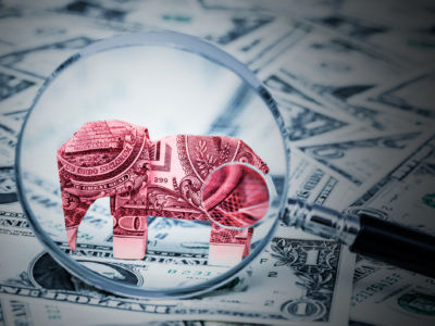 An oragami elephant made of money stands among other us currency while being inspected by a microscope