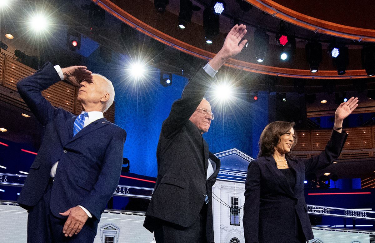 Joe Biden stares into lights as Bernie Sanders and Kamala Harris wave to the audience in front of them