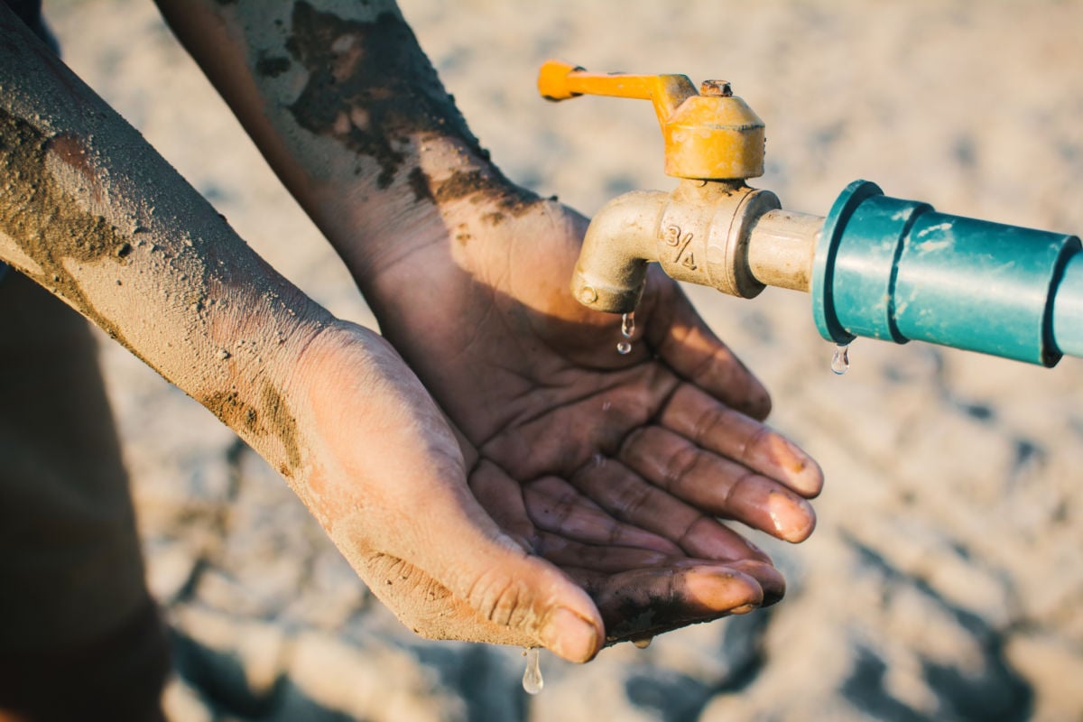 A child's dust-caked hands are cupped underneath a rusted water faucet