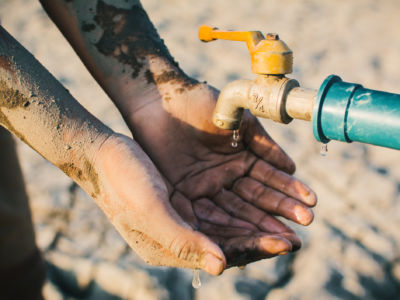 A child's dust-caked hands are cupped underneath a rusted water faucet