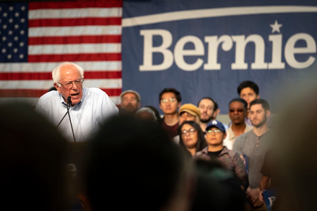 Bernie Sanders speaks at a podium while surrounded by supporters at a rally