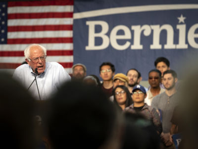 Bernie Sanders speaks at a podium while surrounded by supporters at a rally