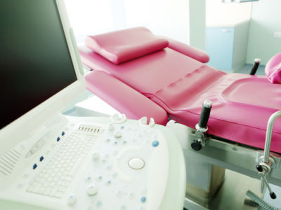 A pink examination table with stirrups sits near a computer monitor