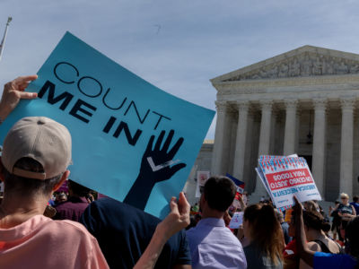 A person holds a blue sign reading "COUNT ME IN" in front of the U.S. Supreme Court building