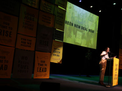 AOC speaks into a mircophone in front of yellow sineage reading "GOOD JOBS AND A LIVABLE FUTURE"