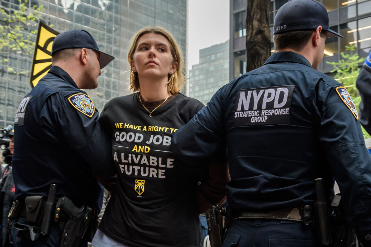 A protester wearing a shirt reading "GOOD JOBS AND A LIVABLE FUTURE" is carried away by cops