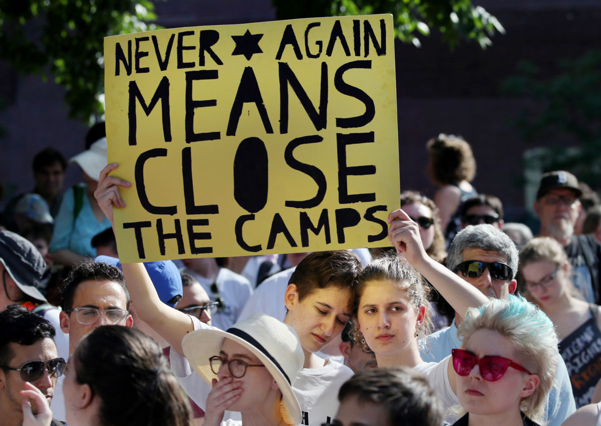 A protester holds a yellow sign reading "NEVER AGAIN MEANS CLOSE THE CAMPS" while surrounded by other activists