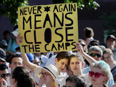 A protester holds a yellow sign reading "NEVER AGAIN MEANS CLOSE THE CAMPS" while surrounded by other activists