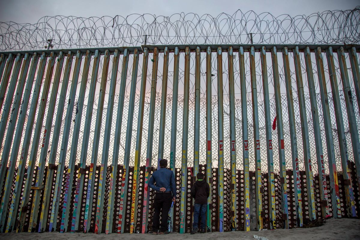 A man and a child look through the slats of the pre-existing border fence along the U.S. / Mexico border