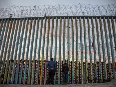 A man and a child look through the slats of the pre-existing border fence along the U.S. / Mexico border