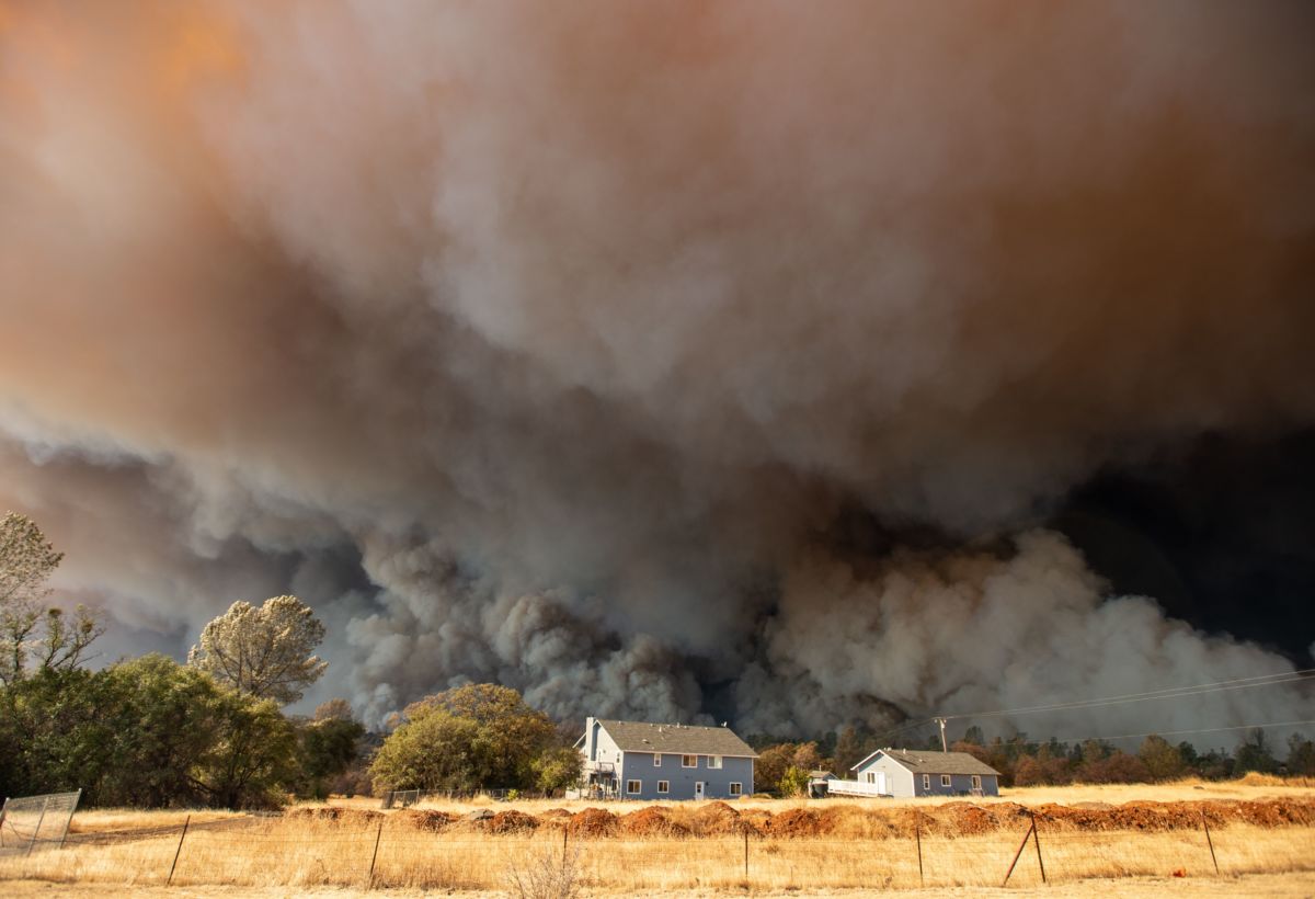 A house is seen in the middle distance as huge plumes of smoke from a wildfire rise behind it