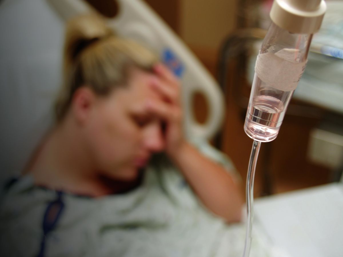 A woman in a hospital bed cradles her head as an iv drip is seen in the foreground