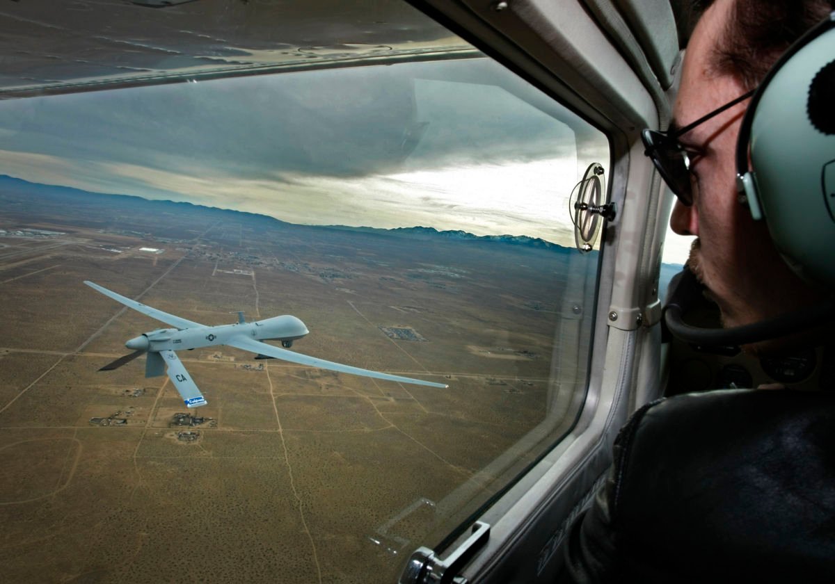 A man in the cockpit of an aircraft looks out of the window at a predator drone flying below him