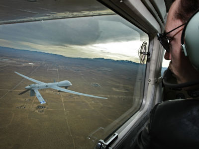 A man in the cockpit of an aircraft looks out of the window at a predator drone flying below him