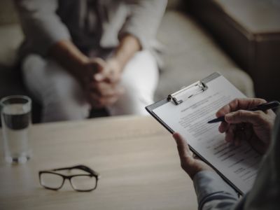 A patient sits in a chair as a doctor looks over forms on a clipboard