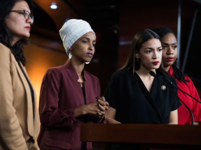 Rashida Tlaib, Ilhan Omar, Alexandria Ocasio-Cortez and Ayanna Pressley look towards the press while standing together at a podium