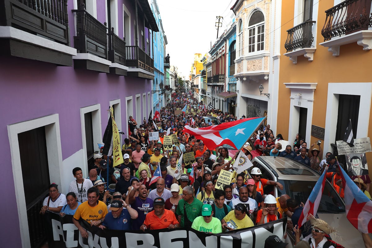 A crowd of people protest while waving Puerto Rican flags and holding signs