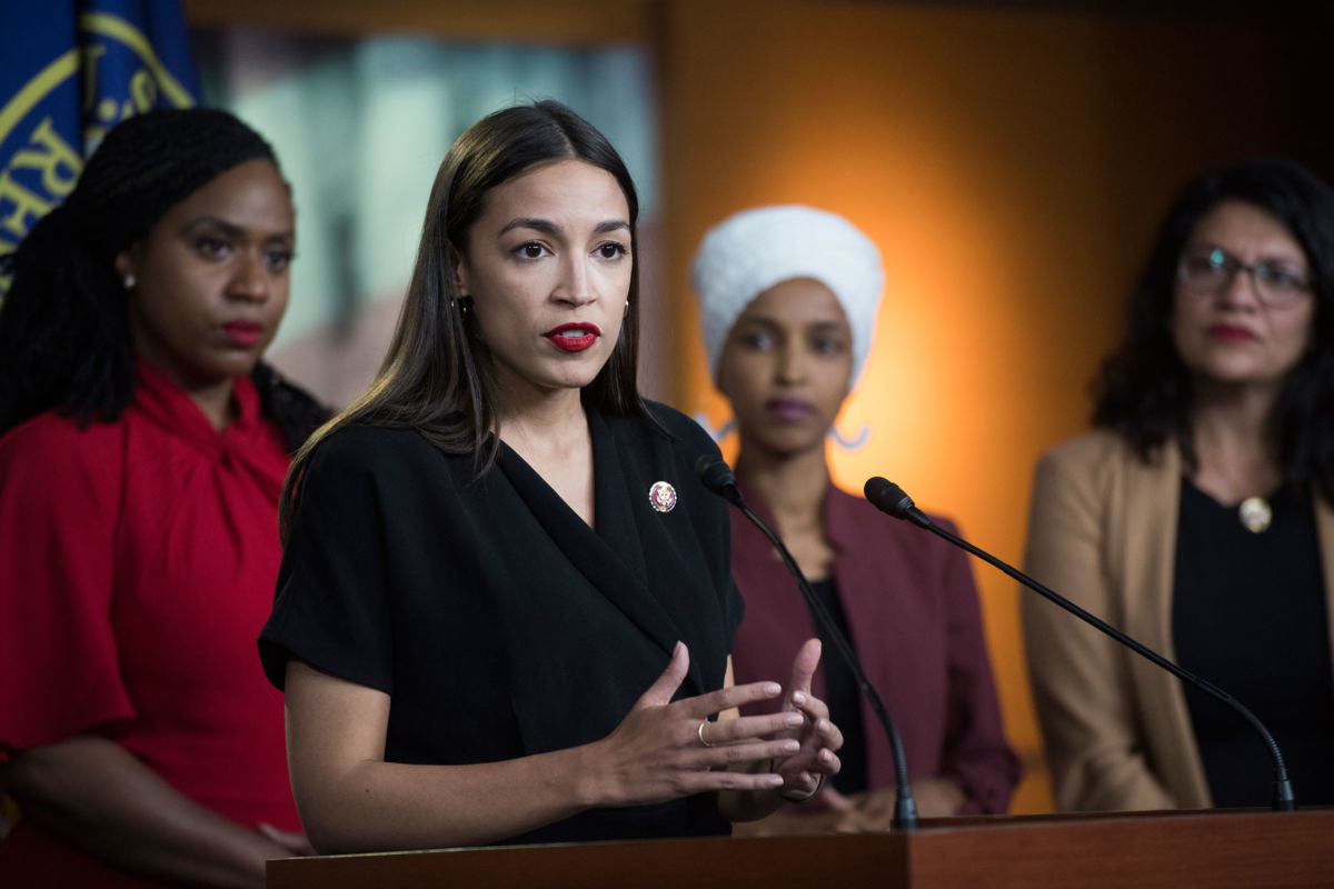 Rashida Tlaib, Ilhan Omar, Alexandria Ocasio-Cortez and Ayanna Pressley look towards the press while standing together at a podium