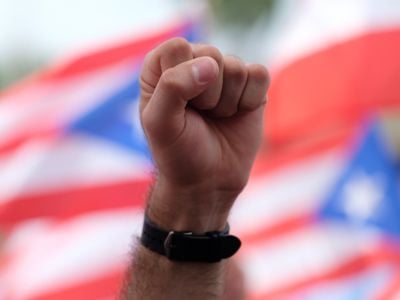 A demonstrator raises a fist during a protest with Puerto Rican flags billowing behind hin