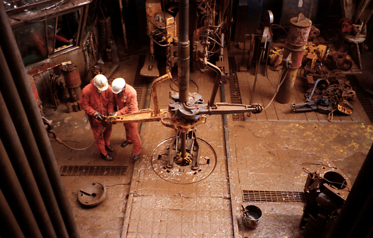 Two workers in orange jumpsuits work in an oil-slicked industrial setting