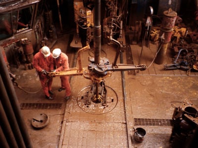 Two workers in orange jumpsuits work in an oil-slicked industrial setting