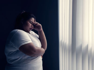 An exhausted-looking woman stands against a grey wall in front of a window