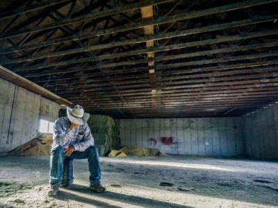 A man in a cowboy hat sits on a bucket in a nearly empty barn and cradles his face in his hands