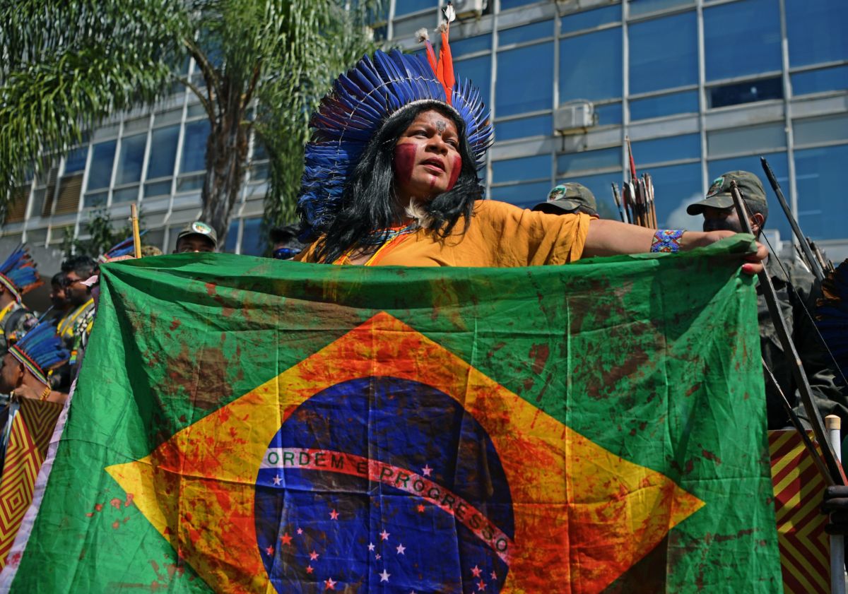 A woman in a headdress holds a Brazilian flag splattered with red paint to simulate bloodstains