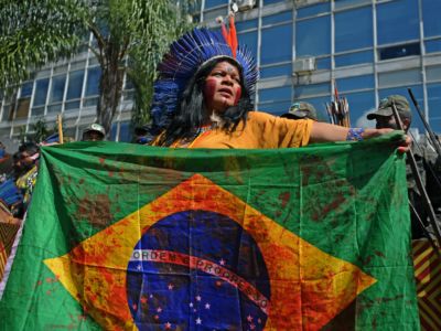 A woman in a headdress holds a Brazilian flag splattered with red paint to simulate bloodstains