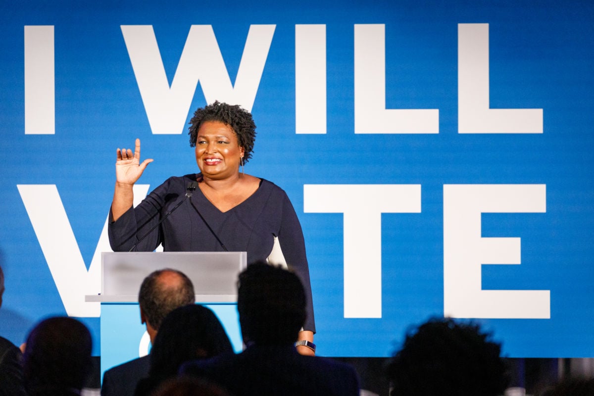 Stacey Abrams stands in front of a blue banner with white text reading "I WILL VOTE"