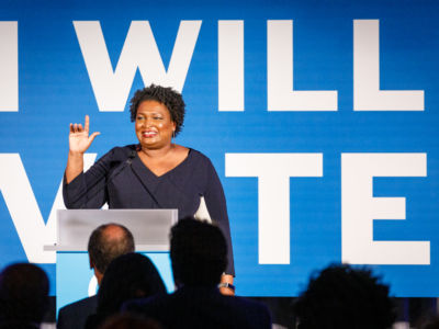 Stacey Abrams stands in front of a blue banner with white text reading "I WILL VOTE"