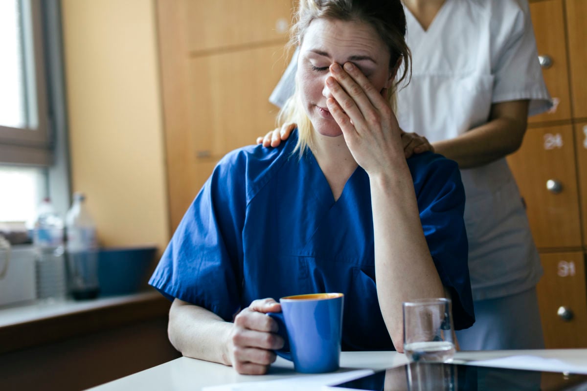 An exhausted nurse holds a cup of coffie while being comforted by a coworker