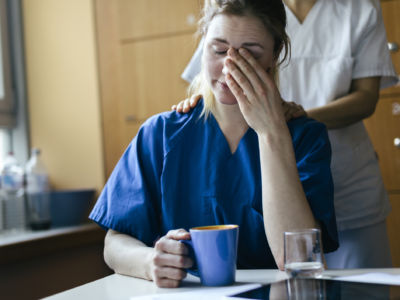 An exhausted nurse holds a cup of coffie while being comforted by a coworker