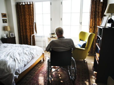 An elderly man sits in a wheelchair in a bedroom