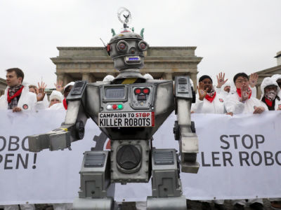 Activists clad in white clothing stand behind a robot puppet with a sign reading "STOP KILLER ROBOTS"