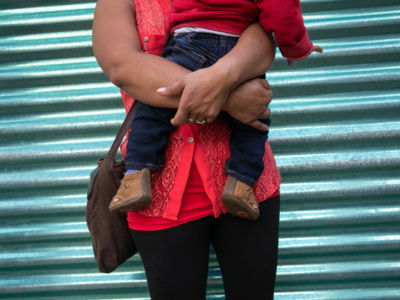 A mother holds her child while standing in front of a corrugated metal fence