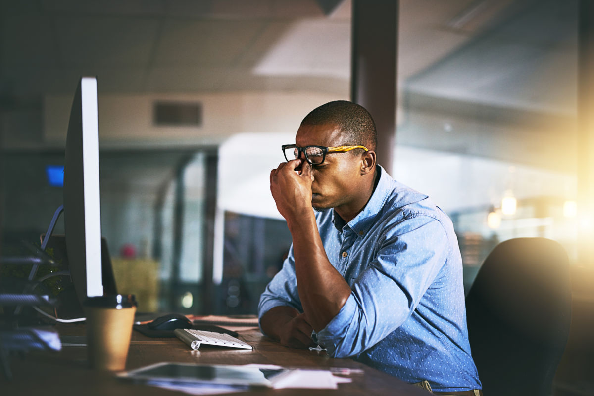 A man sits in front of a computer at a desk and pinches the bridge of his nose