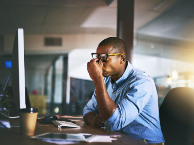 A man sits in front of a computer at a desk and pinches the bridge of his nose