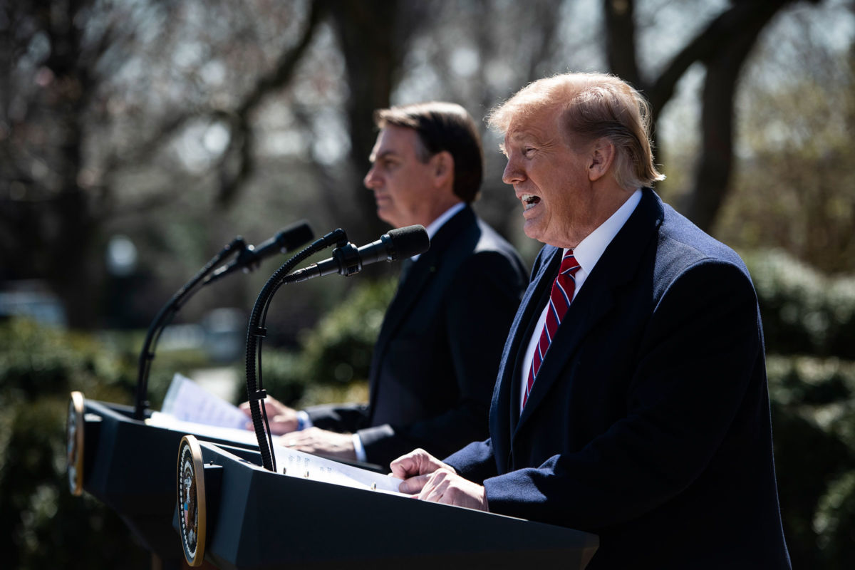 Donald Trump yells into a microphone while Jair Bolsonaro stands at a podium next to him