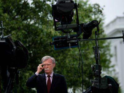 John Bolton adjusts his glasses in front of microphones while being questioned by the press