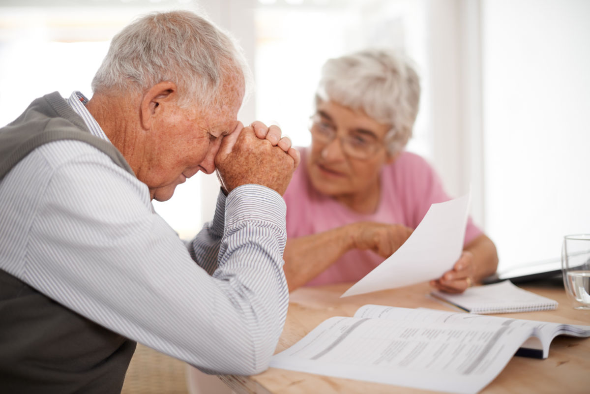 An elderly woman speaks to her husband about the bills in front of them as he rests his temple against his hands