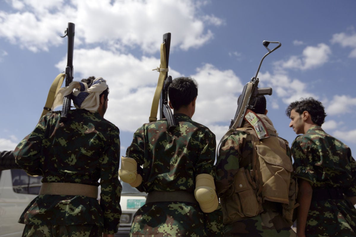 Four soldiers with guns slung over their shoulders stand with their backs to the camera