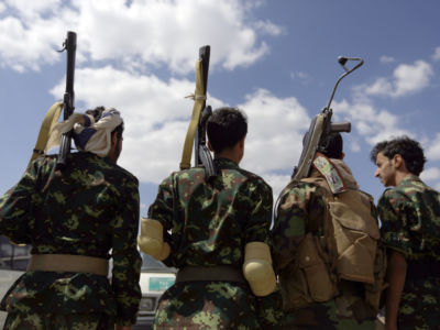 Four soldiers with guns slung over their shoulders stand with their backs to the camera