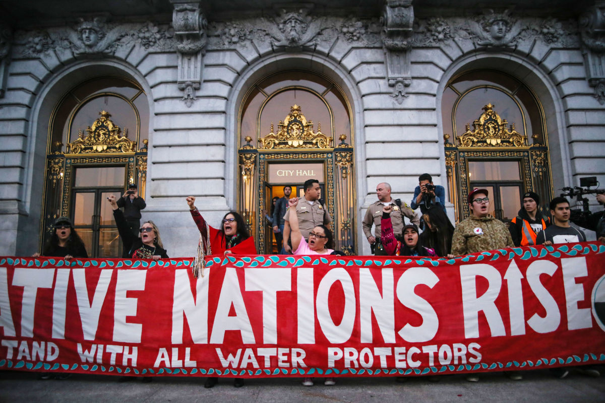 Water Protectors stand behind a red sign reading "NATIVE NATIONS RISE"