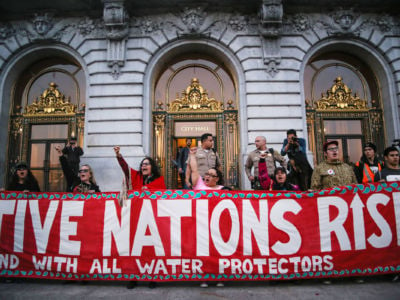 Water Protectors stand behind a red sign reading "NATIVE NATIONS RISE"