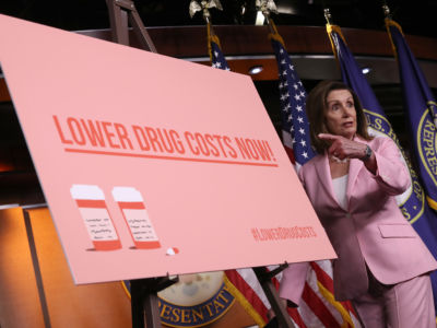 Nancy Pelosi gestures toward a pink sign reading "LOWER DRUG COSTS NOW!"
