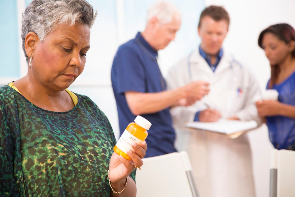 A woman looks at a bottle of pills in her hand as her medical providers talk with eachother in the background