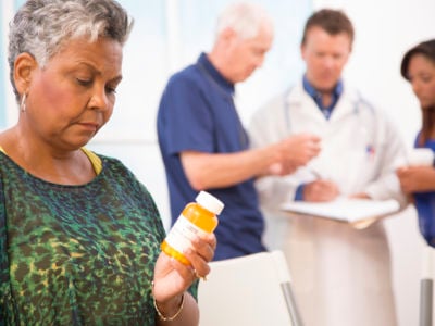 A woman looks at a bottle of pills in her hand as her medical providers talk with eachother in the background